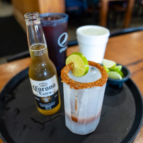 Corona beer bottle, michelada with lime, and other drinks on a black tray.