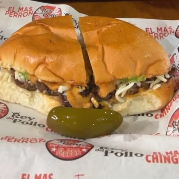 A Rey del Pollo Mexican sandwich, cut in half, with a green pepper on branded paper.