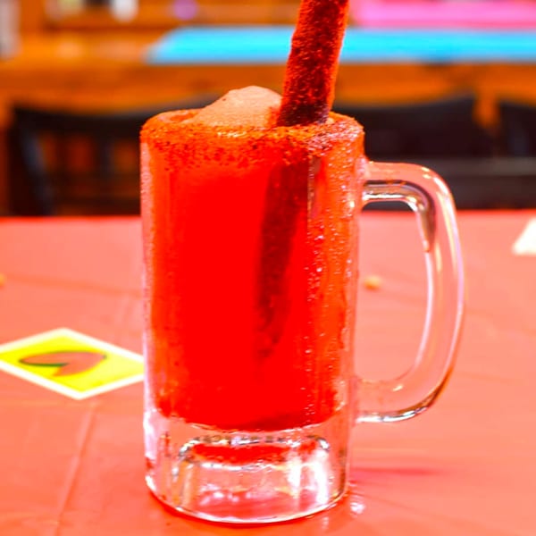 A vibrant red Mexican drink in a glass mug, rimmed with chili powder and a tamarind candy straw.