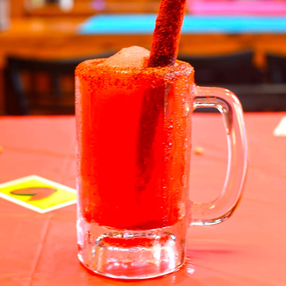 A vibrant red Mexican drink in a glass mug, rimmed with chili powder and a tamarind candy straw.